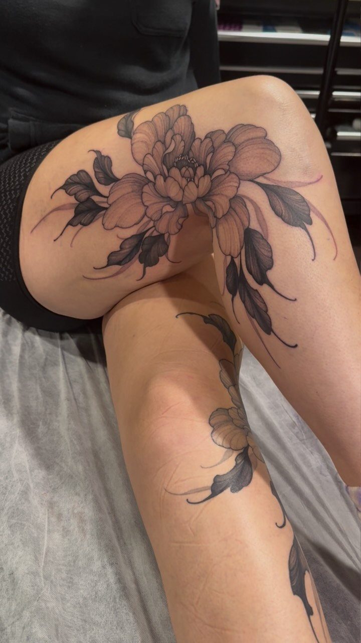 Large black and grey peony tattoo with shaded petals on a person's thigh and leg.