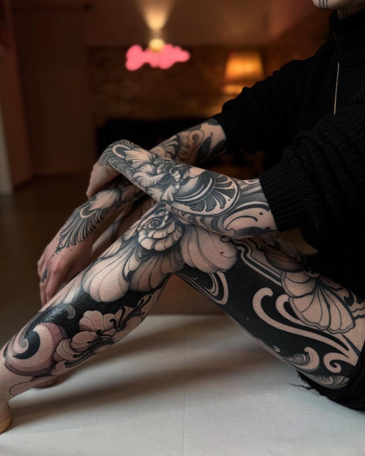 Large floral blackwork tattoos covering a person's arms and thighs while they sit down.