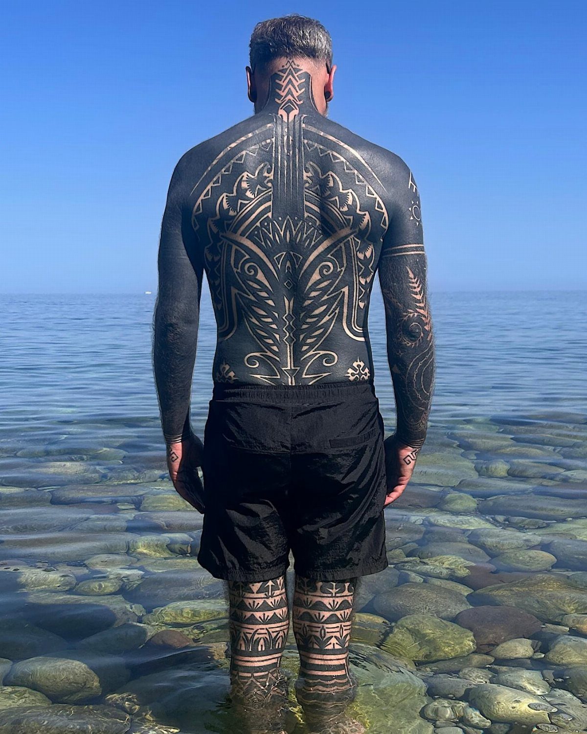 Man standing in clear ocean water showing large tribal blackwork tattoos on his back and legs.