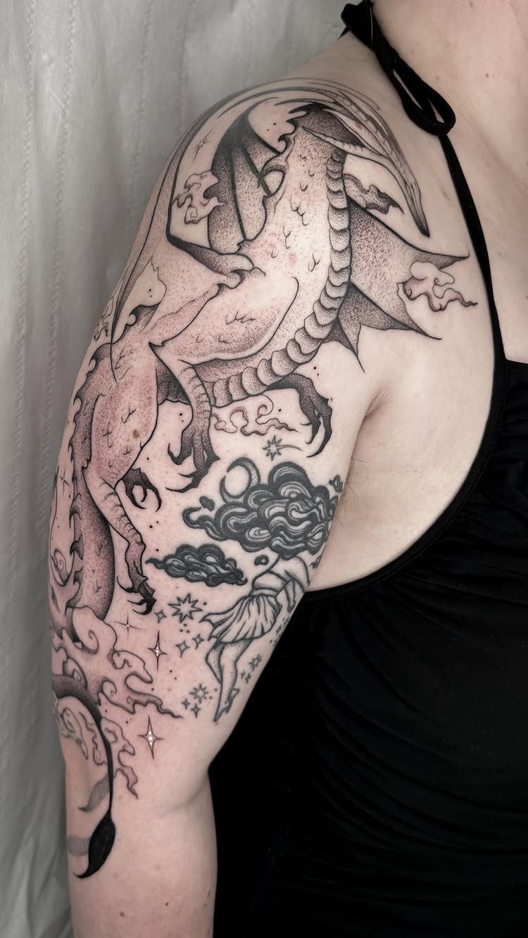 A large illustrative blackwork dragon tattoo with clouds and stars on a person's shoulder.