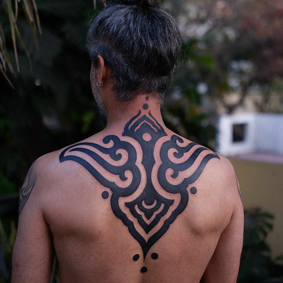 A man's back displaying a large, symmetrical blackwork tattoo with bold tribal patterns.