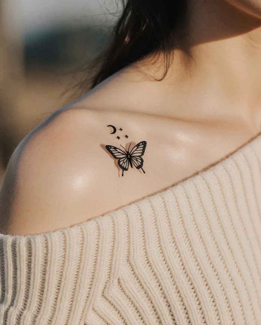 A small black ink butterfly tattoo with stars and a moon on a person's collarbone.