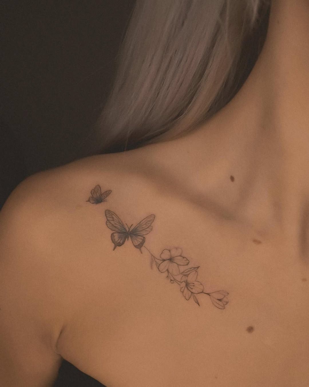 Two delicate black ink butterfly tattoos and a floral branch on a person's collarbone.