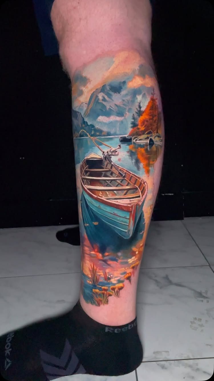 8 Color Tattoo Ideas for Men: Comic, Dragon & Watercolor 5 A colorful landscape tattoo on a leg depicting a boat on a lake with mountains.
