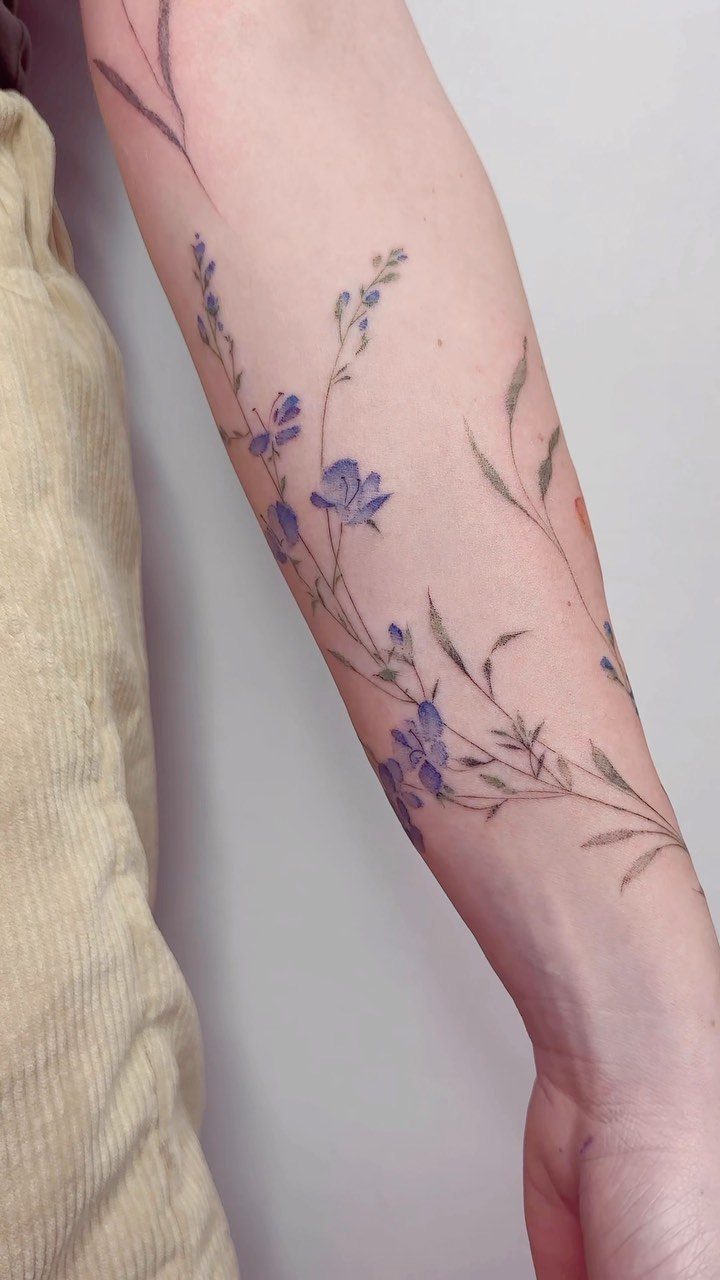 A forearm tattoo of delicate blue wildflowers with thin green stems on pale skin.