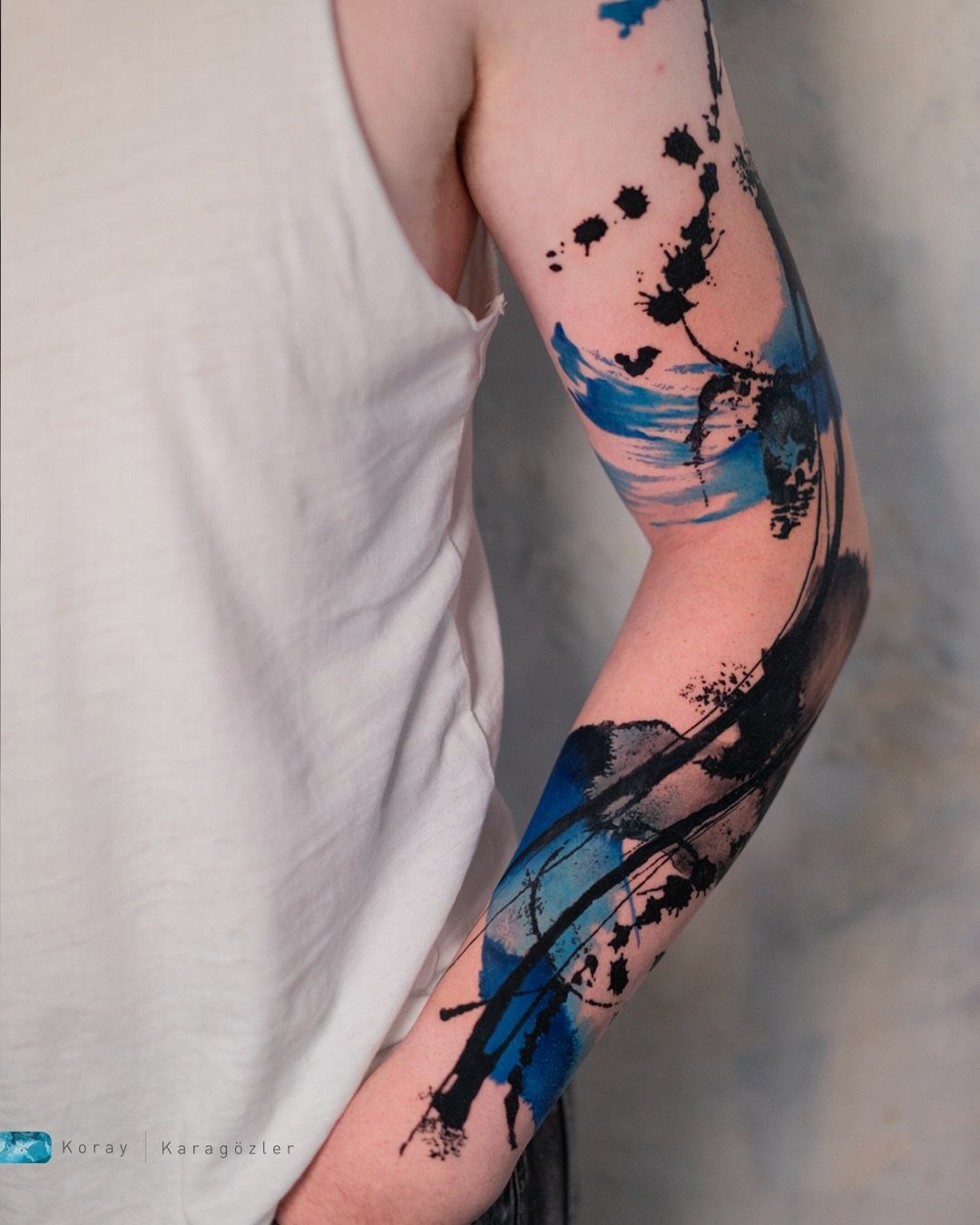 A person's arm displaying an abstract tattoo with black splatters and blue watercolor strokes.
