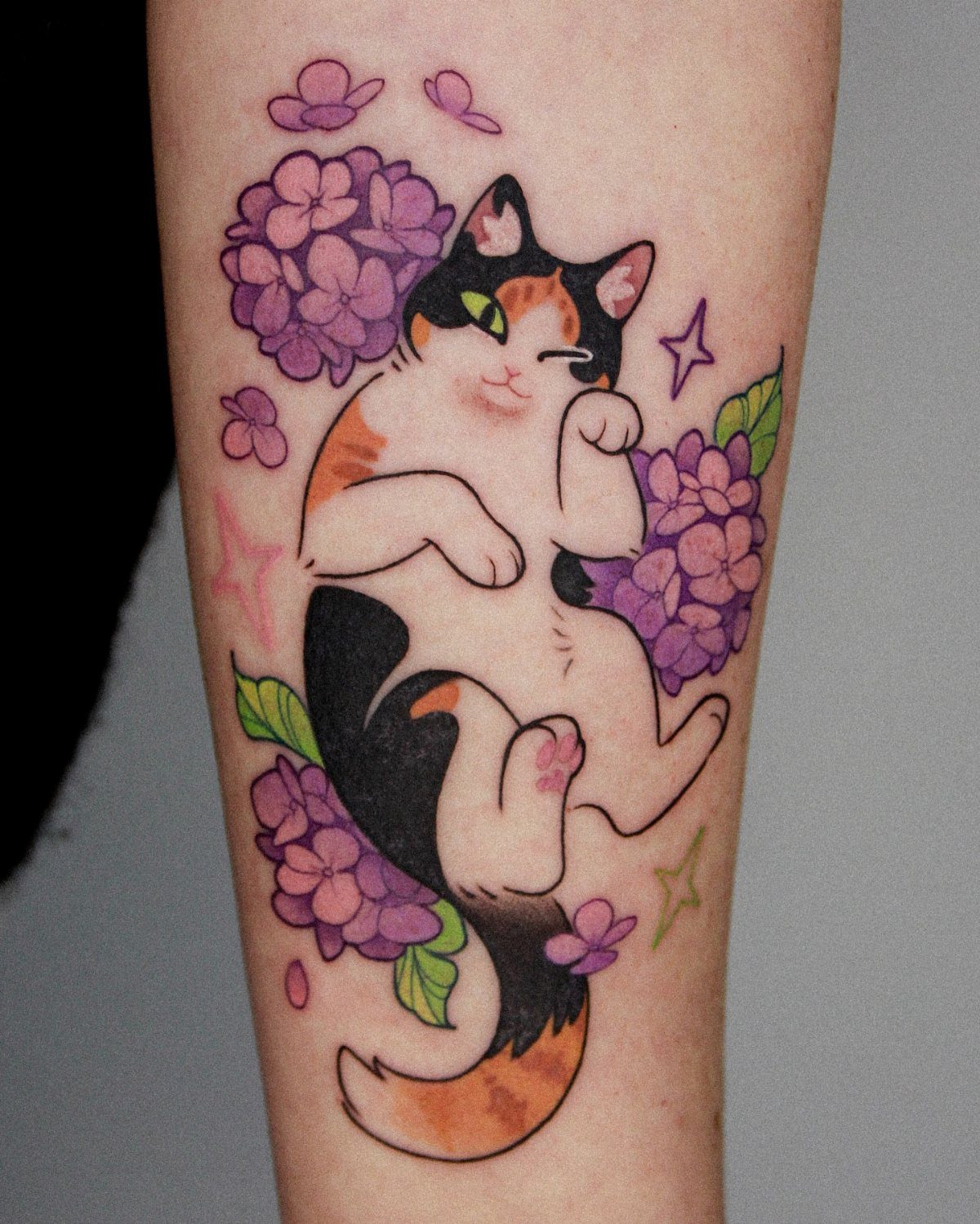 A colorful calico cat tattoo surrounded by purple flowers and pink stars on skin.