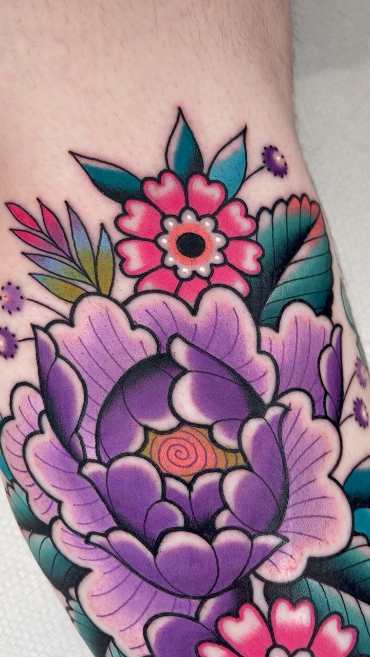 A vibrant purple and pink floral tattoo with teal leaves on a person's skin.
