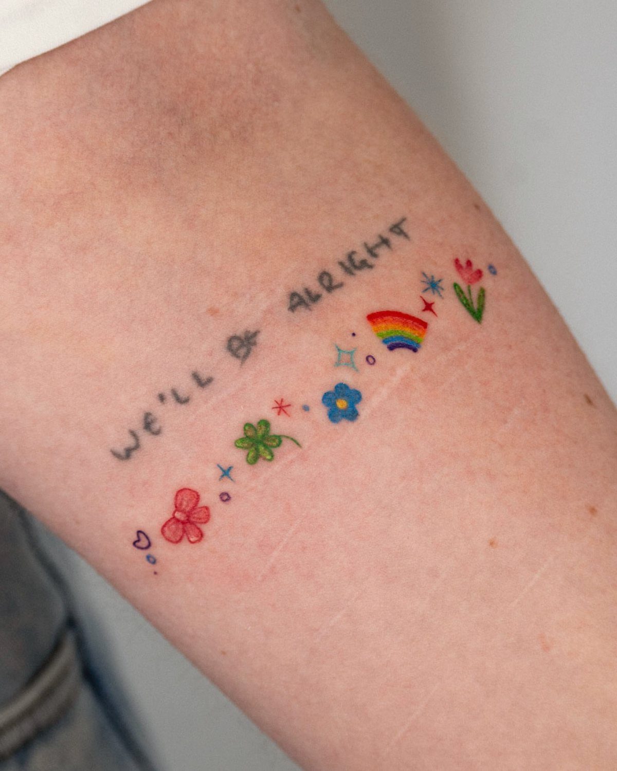 Small colorful tattoo on an arm reading We'll be alright with tiny rainbow and flower icons.