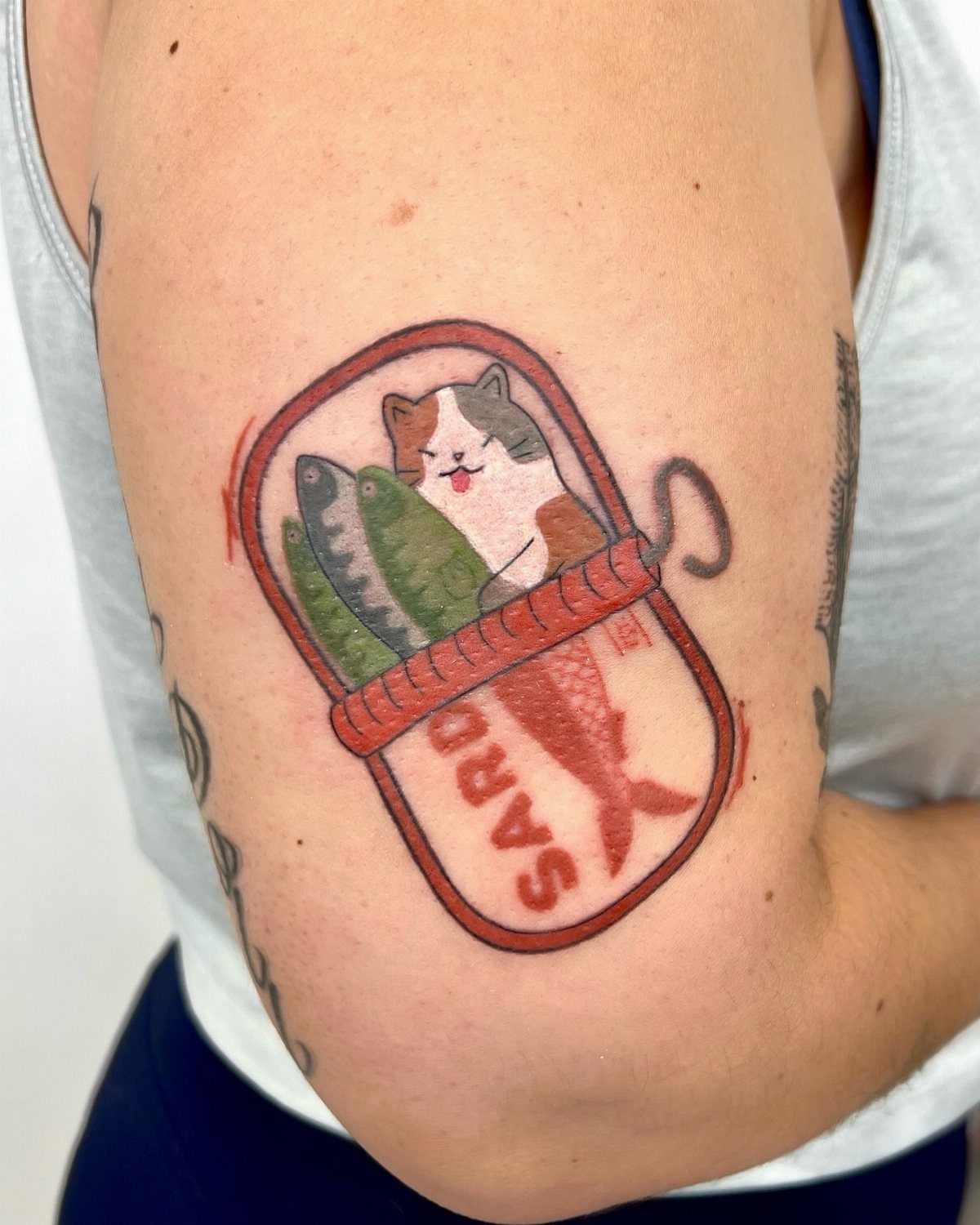 A colorful cartoon cat and fish tattoo inside a sardine tin on a person's arm.