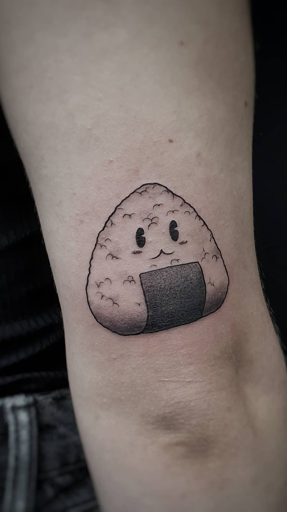 A small black and grey tattoo of a smiling onigiri rice ball on a person's arm.