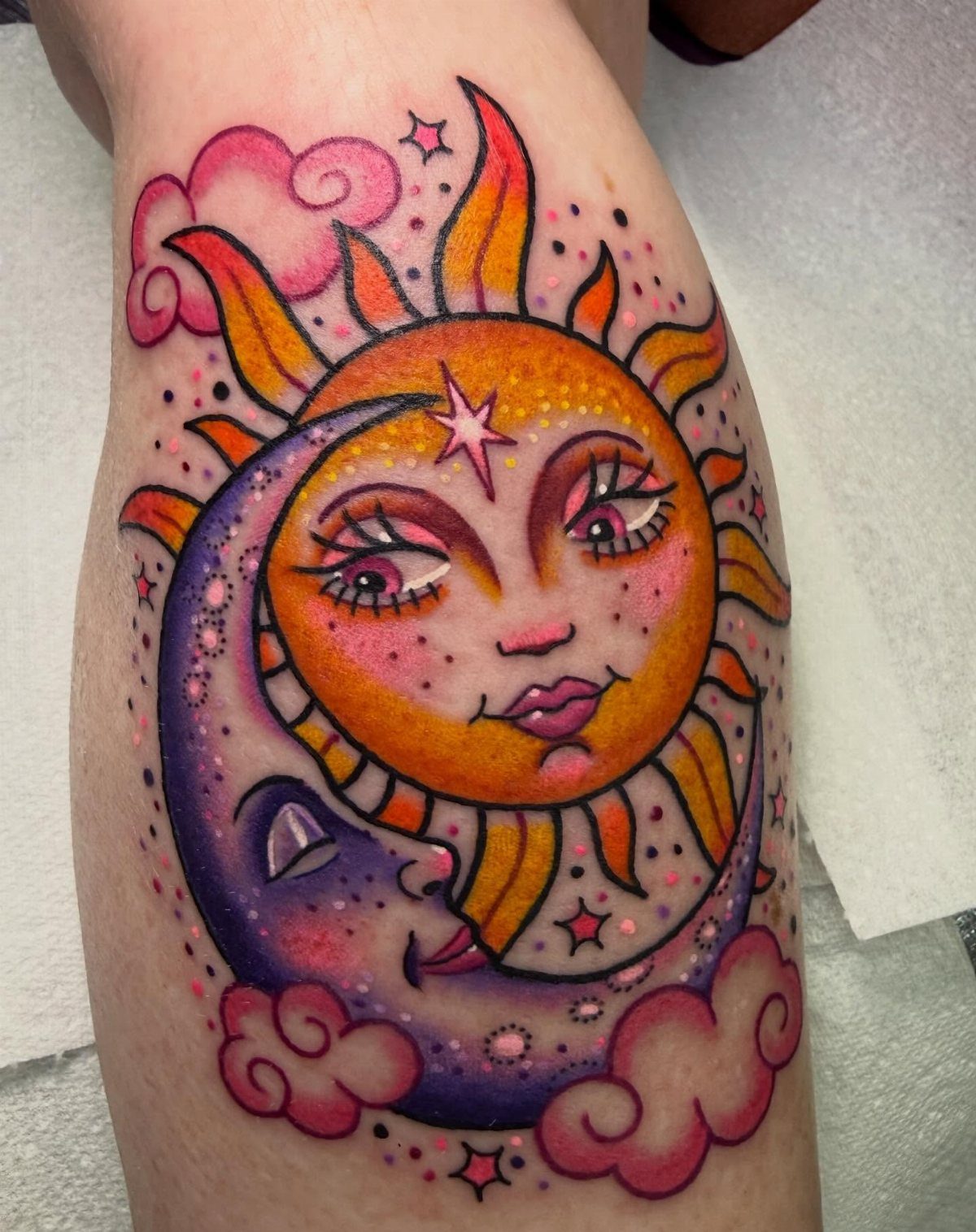 A colorful tattoo of a smiling sun and a purple crescent moon surrounded by pink clouds.