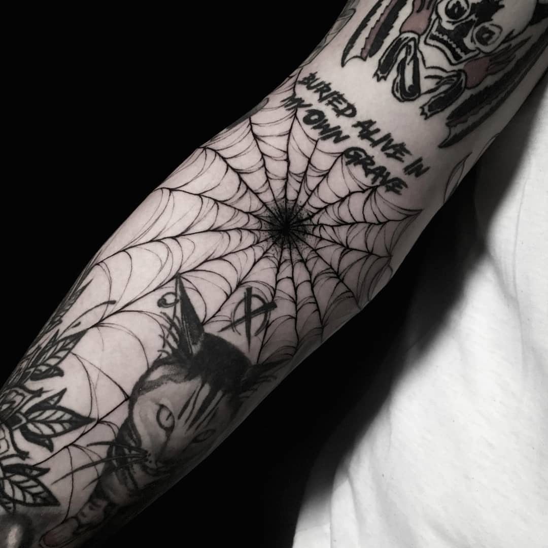 A person's arm covered in various black ink tattoos including a spiderweb and a cat.