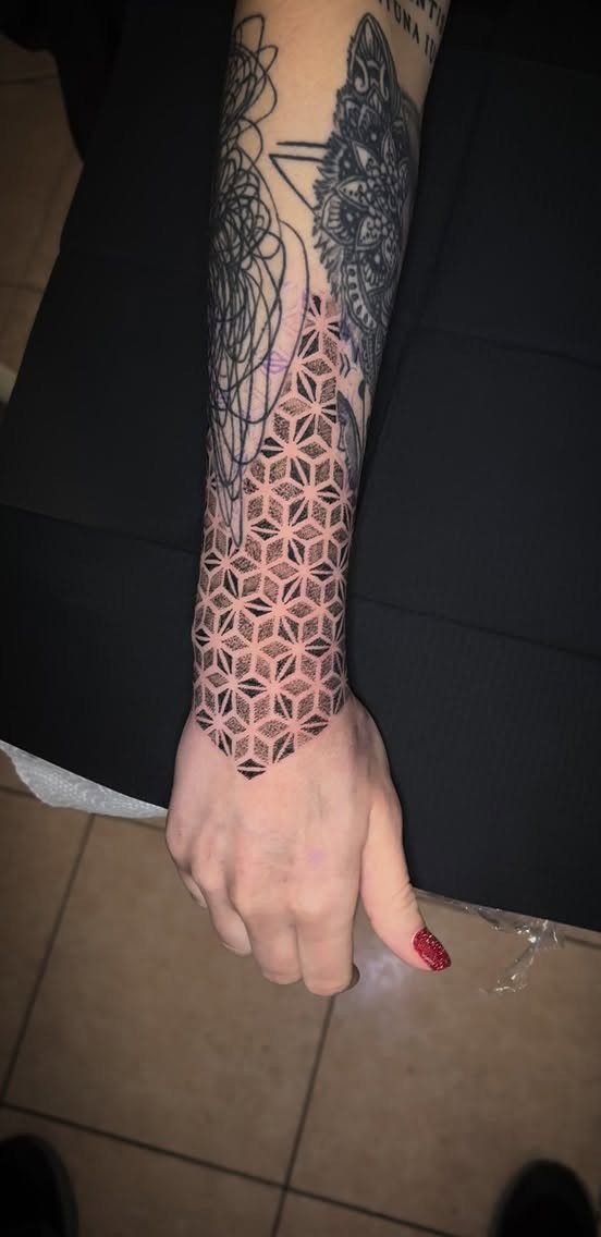 A forearm displaying various black ink tattoos including a prominent geometric pattern of interlocking shapes.