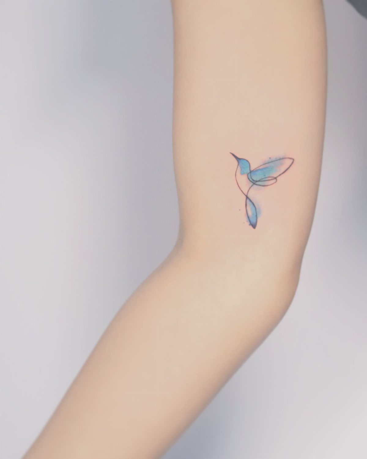 Fine Line Tattoo Aesthetic for Women: 9 Idea Boards 7 A small fine line bird tattoo with blue and pink shading on a person's arm.