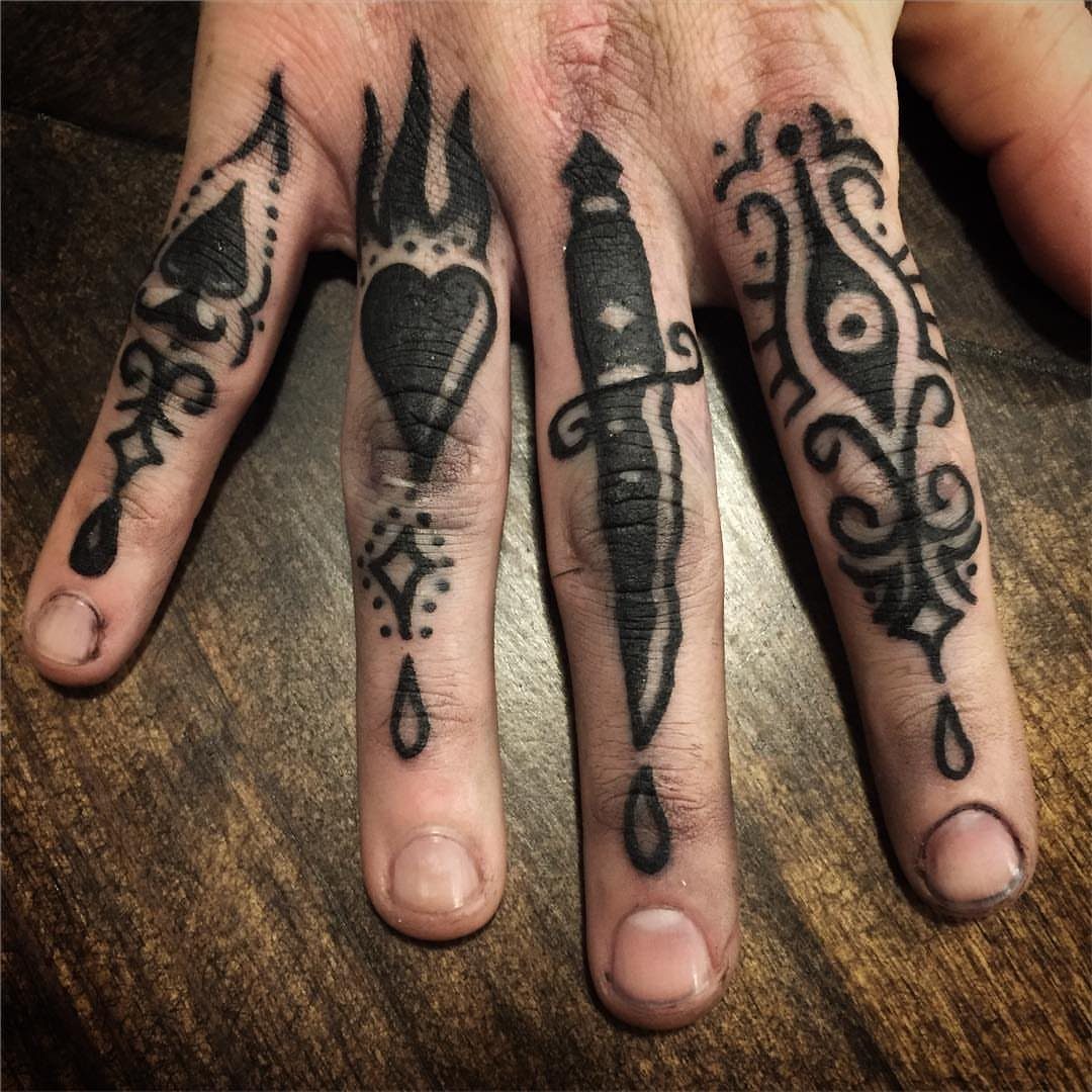Four fingers with black ink tattoos including a spade, heart, dagger, and ornate eye symbol.