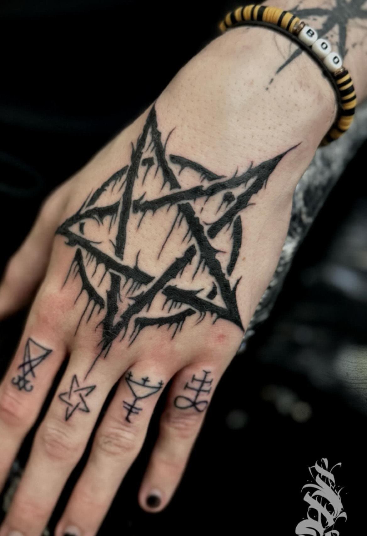 A hand with a large pentagram tattoo and various small occult symbols on the fingers.