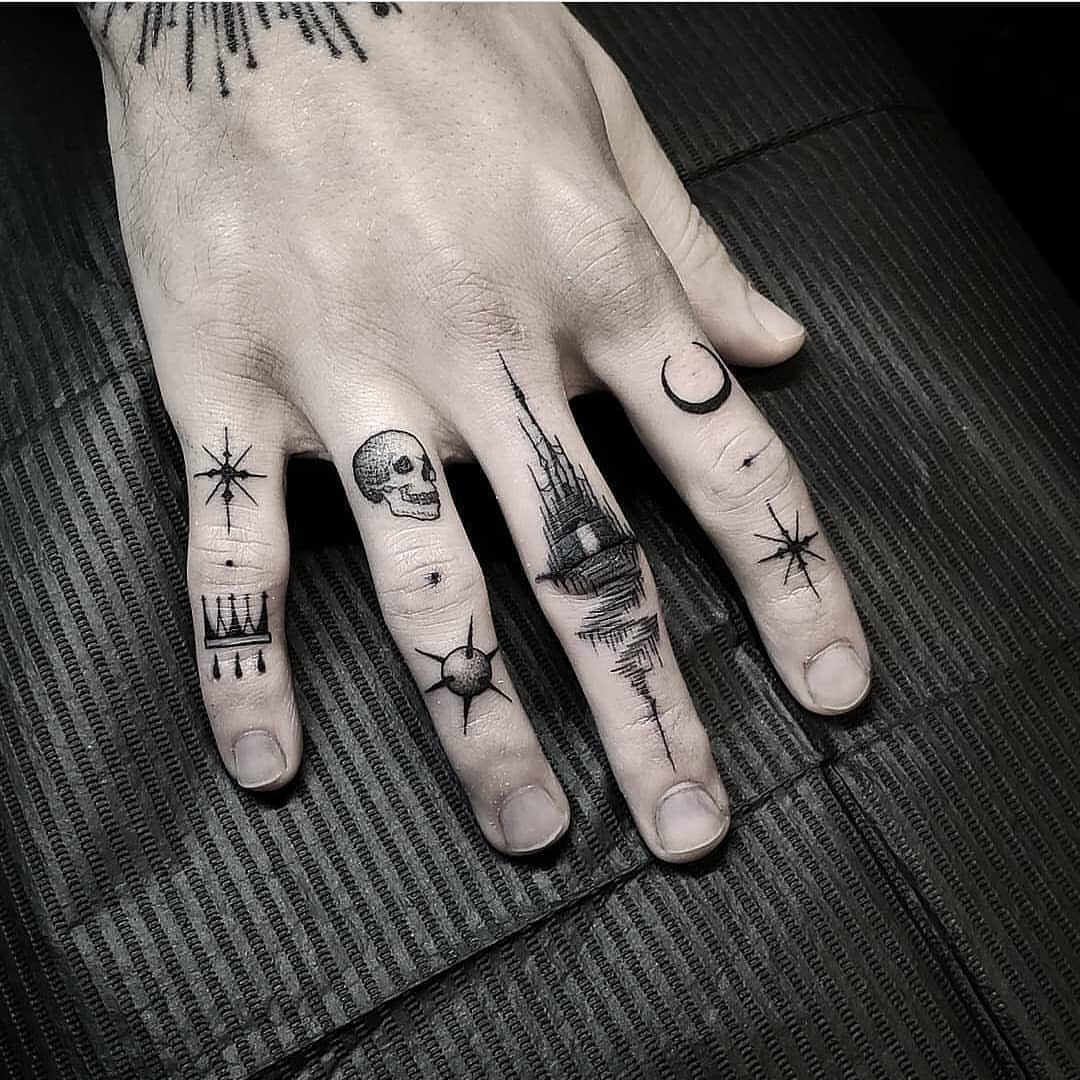 A hand featuring various small black ink finger tattoos including a skull and crescent moon.
