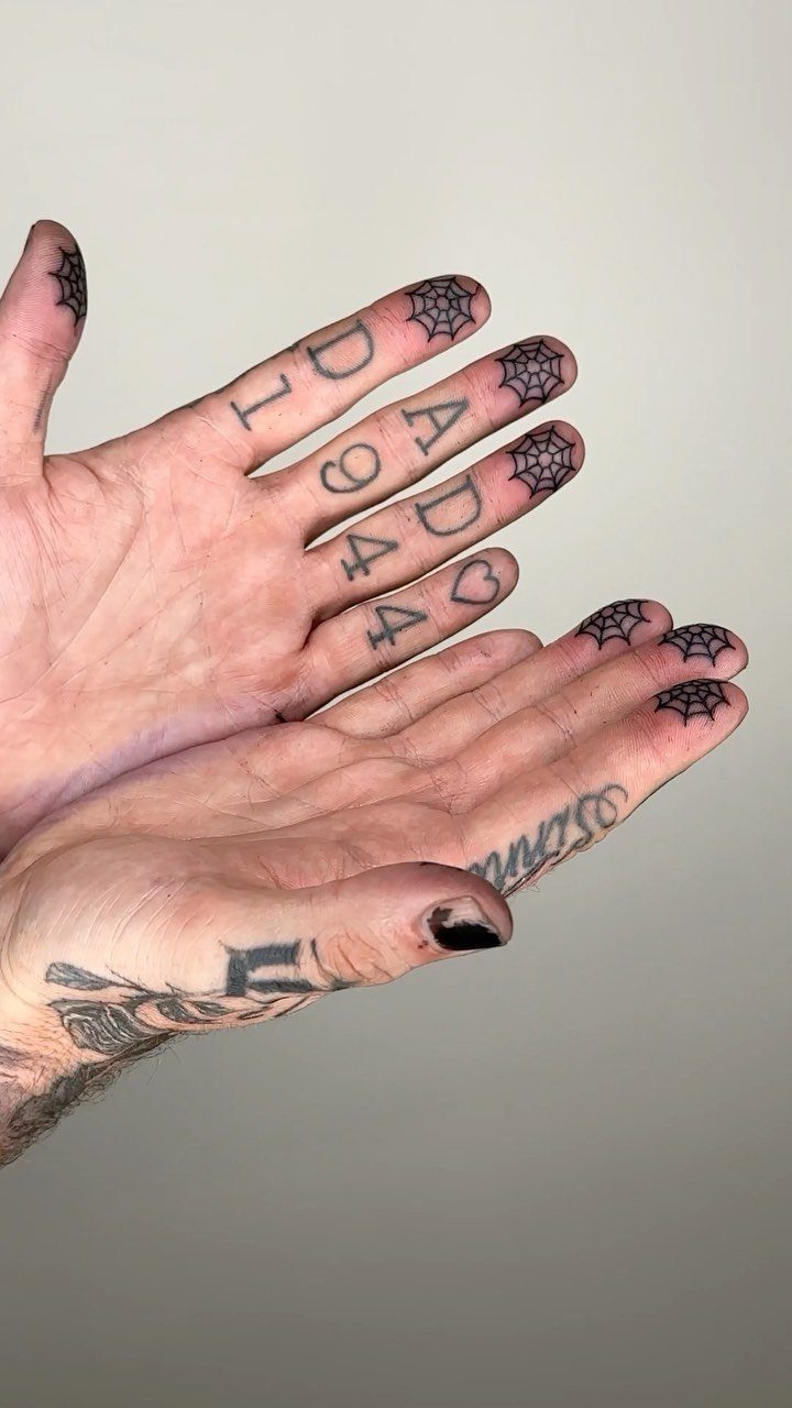 Hands displaying various black ink tattoos including small spiderweb designs on the knuckles.