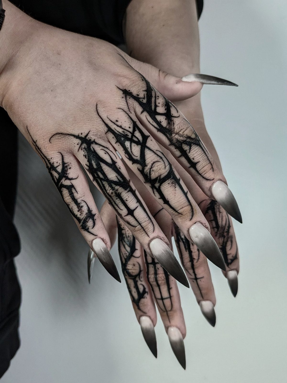 Hands with long pointed nails and black thorny vine tattoos covering the fingers and skin.