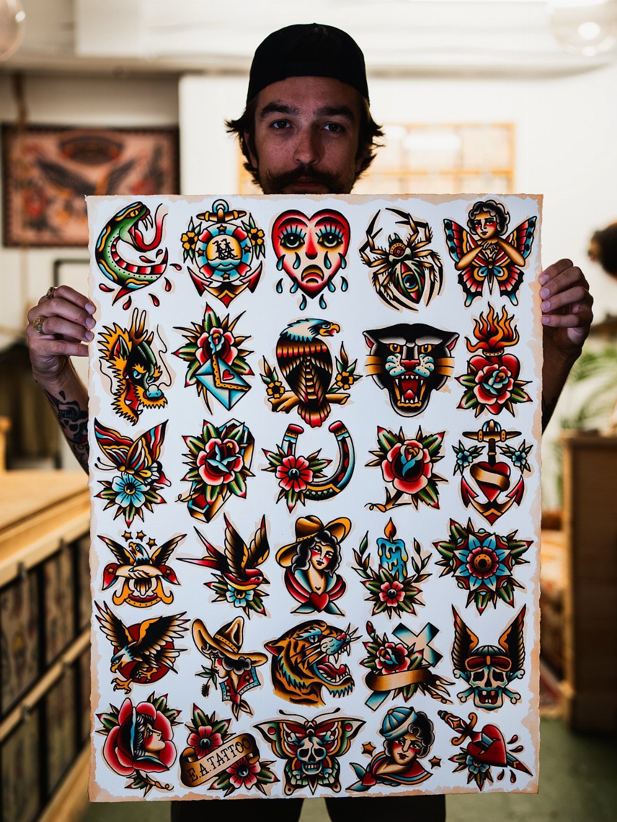 A man holding a large sheet of colorful traditional flash tattoo designs featuring various motifs.