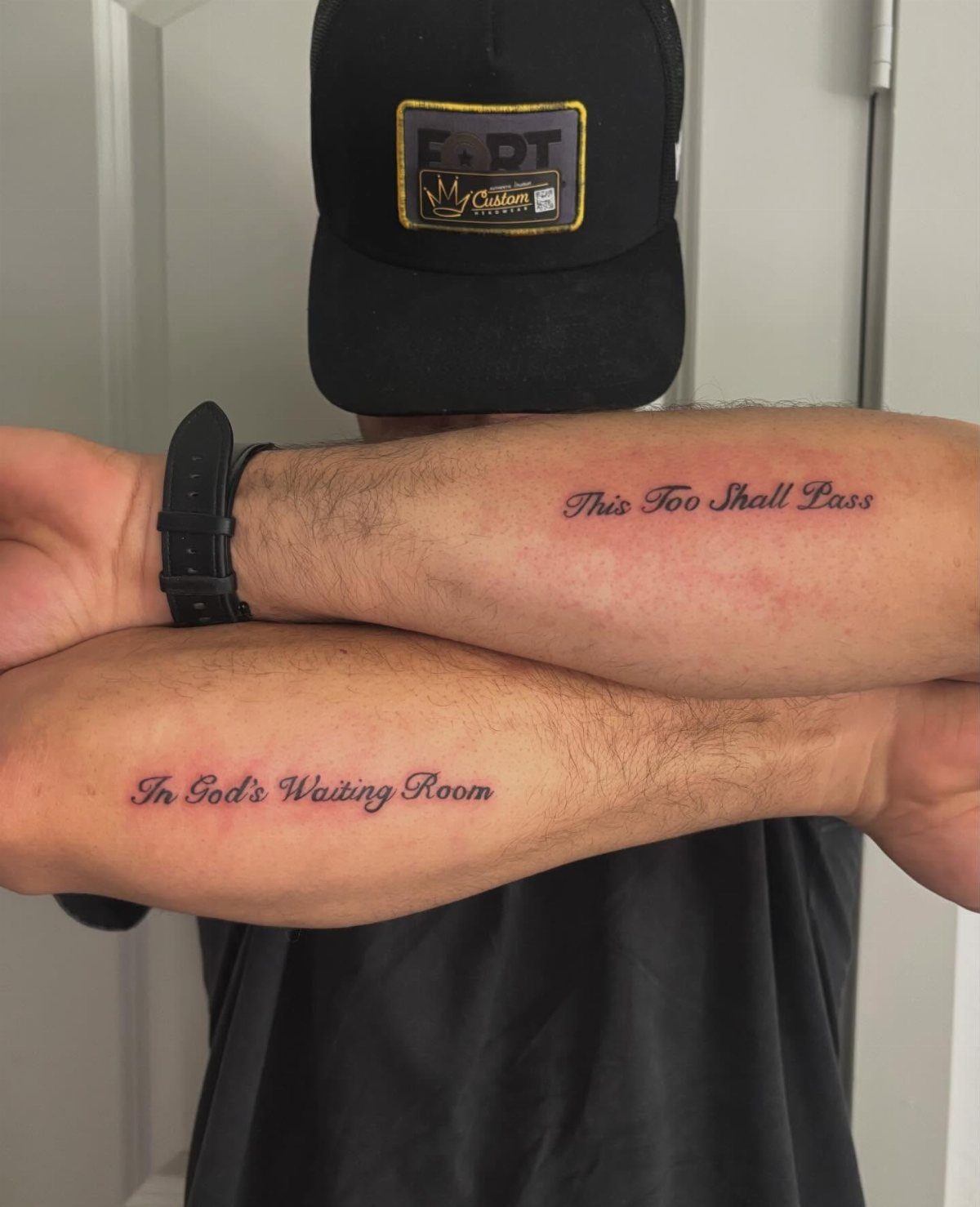 Man's crossed arms displaying black ink script phrase tattoos on the forearms.