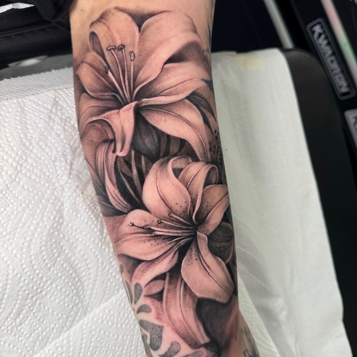 A detailed black and grey realism tattoo of two large lilies on a person's forearm.