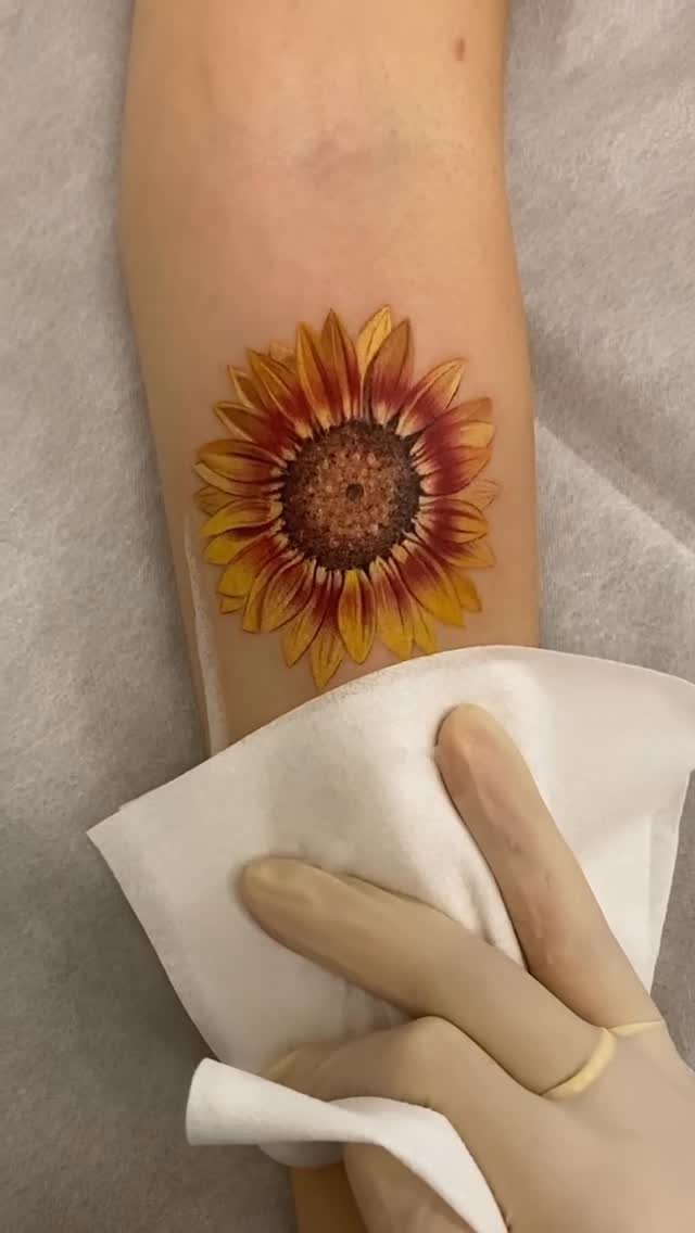 A detailed sunflower tattoo with yellow and orange petals on a person's forearm.