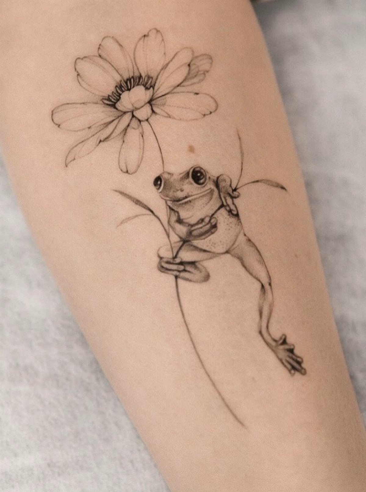 A black and grey fine line tattoo of a frog climbing a daisy stem on skin.