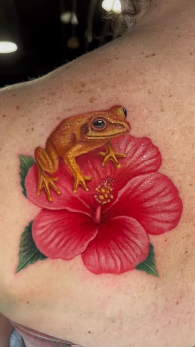 A detailed tattoo of a brown frog sitting on a bright red hibiscus flower.