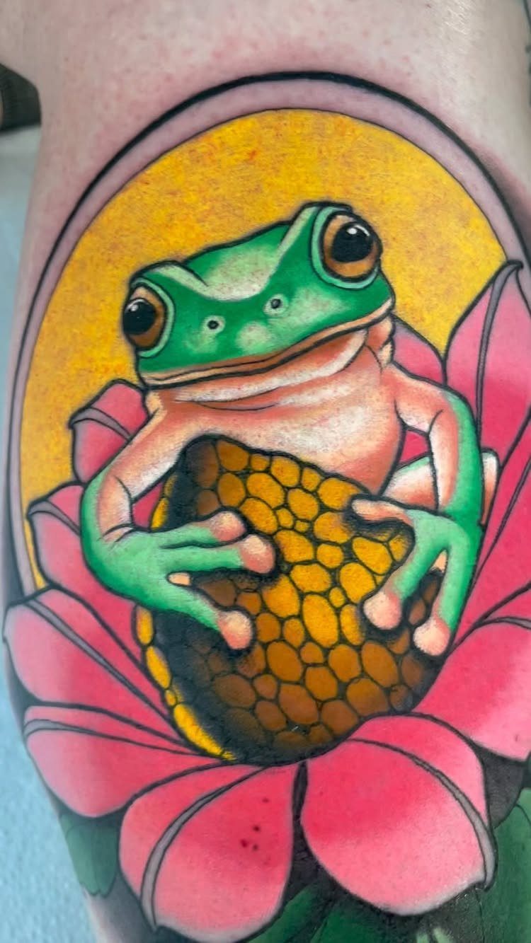 A green tree frog tattoo resting on a vibrant pink lotus flower with a yellow center.