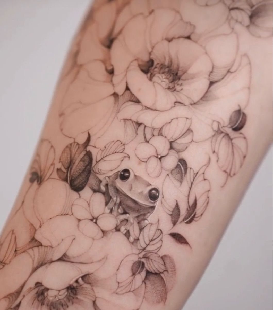 A detailed black and grey tattoo of a small frog surrounded by large peony flowers on skin.