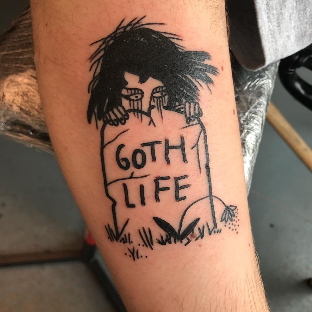 A black ink tattoo on an arm showing a figure behind a tombstone reading GOTH LIFE.