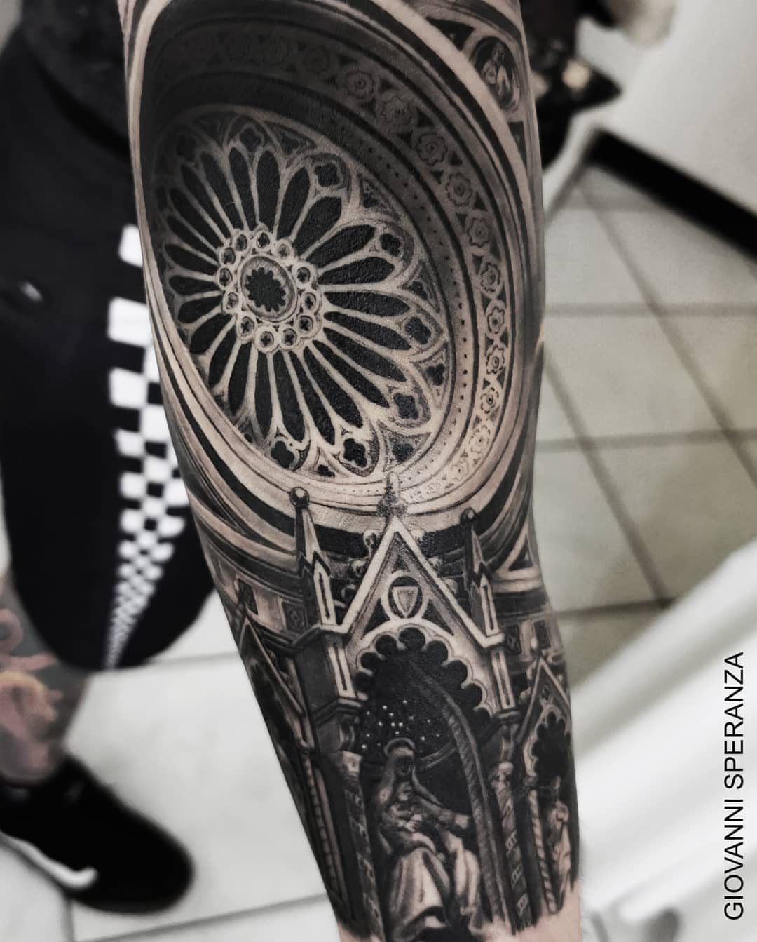 A detailed black and grey tattoo of a Gothic cathedral rose window on a person's arm.