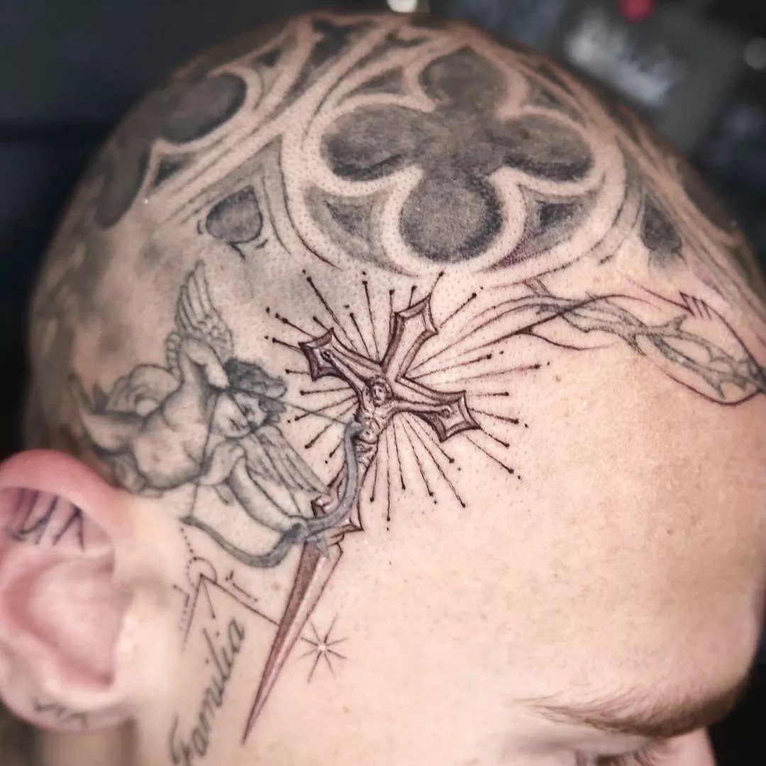 A person's shaved head featuring a detailed gothic crucifix tattoo and an angel tattoo.