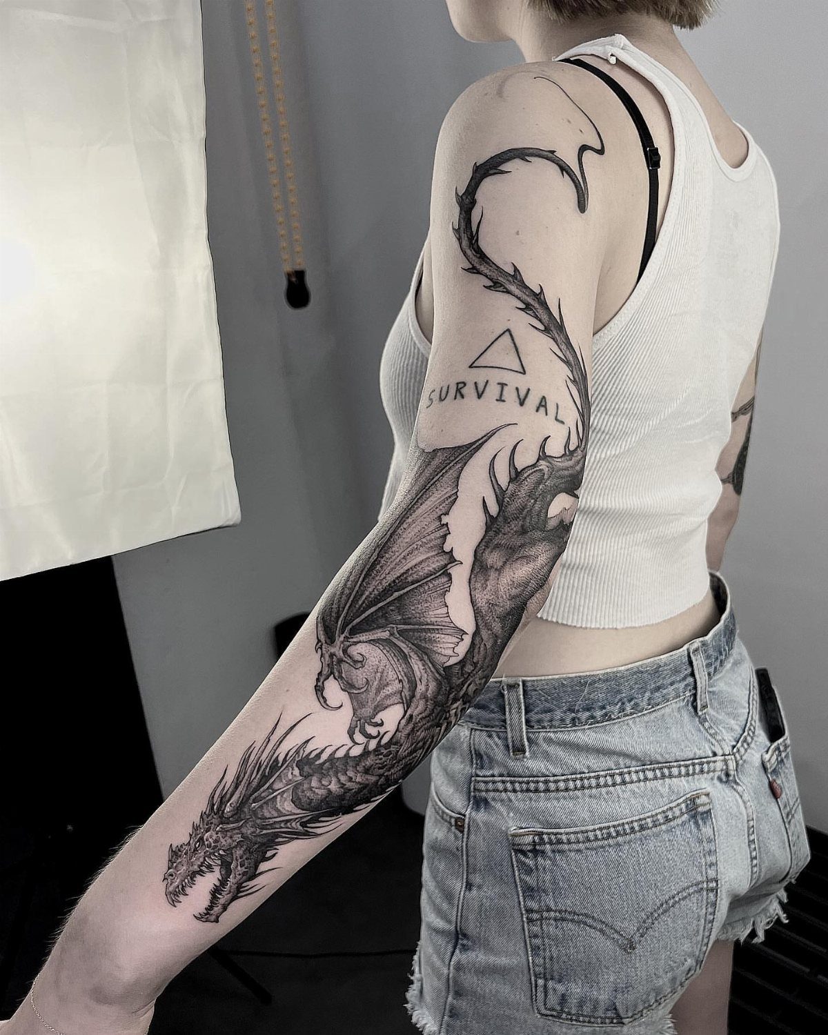 A large black ink dragon tattoo wrapping around a person's arm with the word survival.