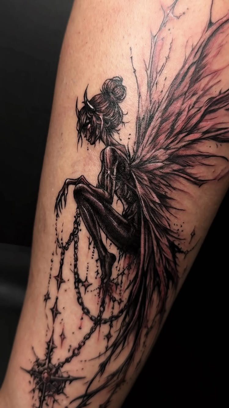 A detailed black and grey gothic fairy tattoo with large wings on a person's arm.