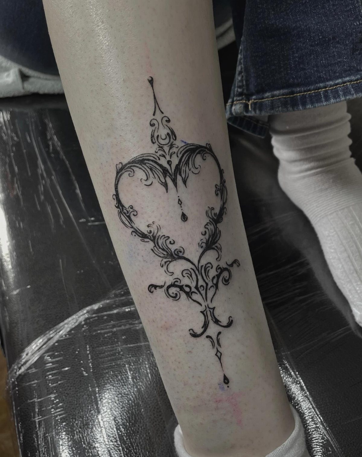 An ornamental black ink heart-shaped tattoo on a person's lower leg.
