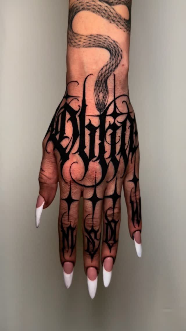 A hand and forearm covered in large, black gothic lettering and snake-like patterned tattoos.
