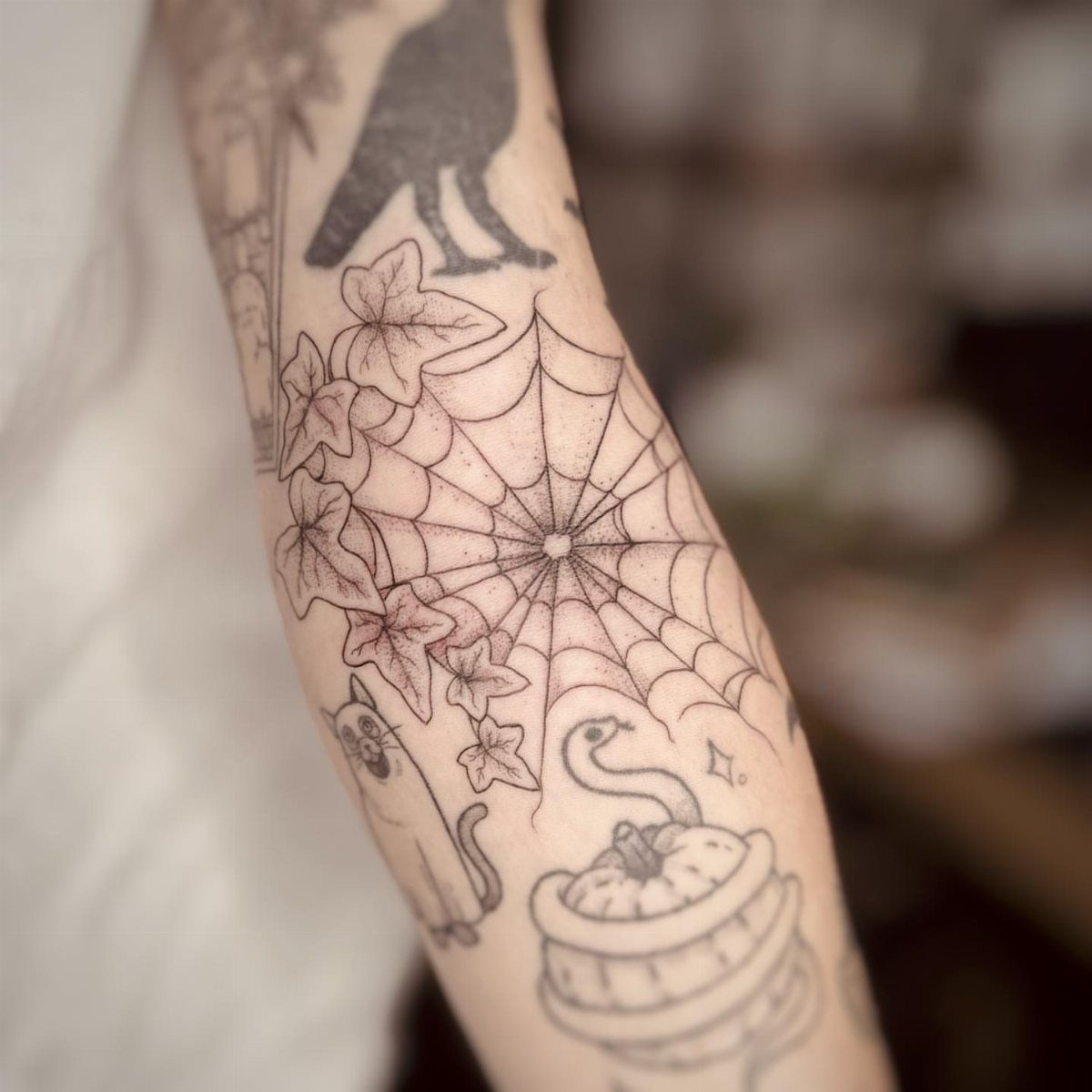 A person's arm decorated with black ink tattoos of a spider web, raven, and ivy leaves.
