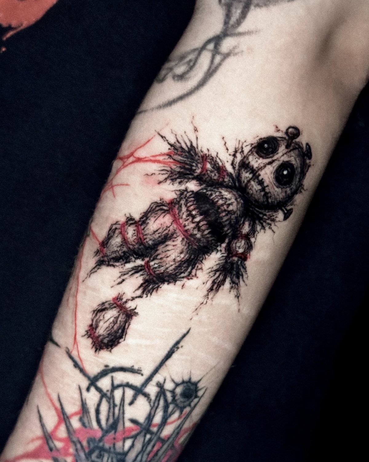 A black and red voodoo doll tattoo on a person's forearm with button eyes.