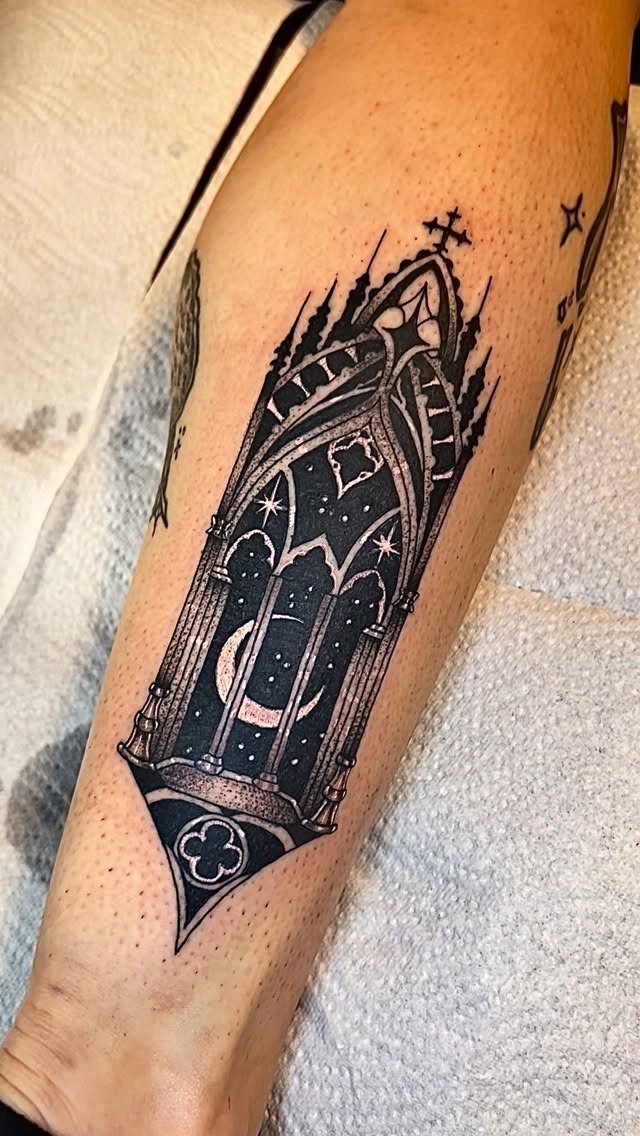 A black ink gothic window tattoo with stars and a crescent moon on a person's leg.