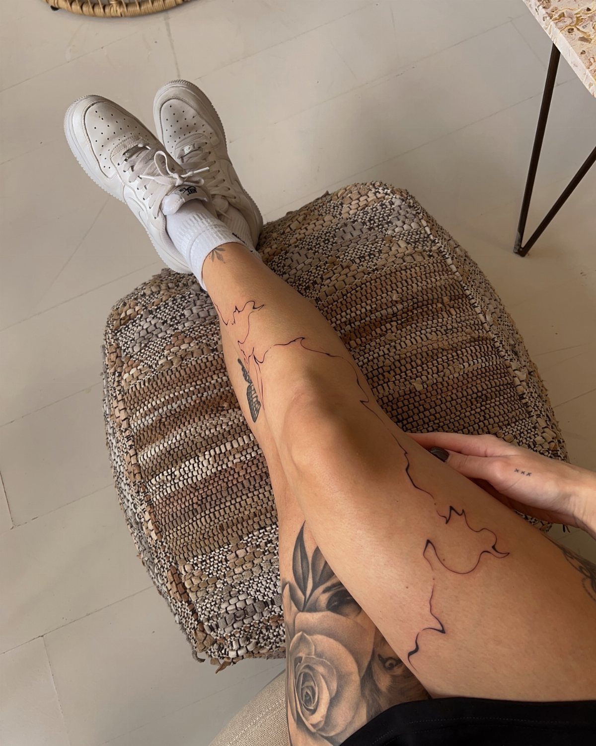 7 Leg Tattoo Ideas for Women: Geometric, Floral, Fine Line & More 3 A person's tattooed legs resting on a woven ottoman in a bright room.