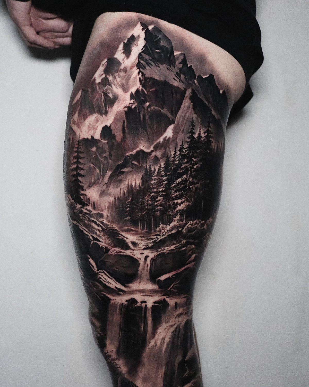7 Leg Tattoo Ideas for Women: Geometric, Floral, Fine Line & More 6 A detailed black and grey mountain landscape tattoo featuring snowy peaks, pine trees, and a waterfall on a thigh.