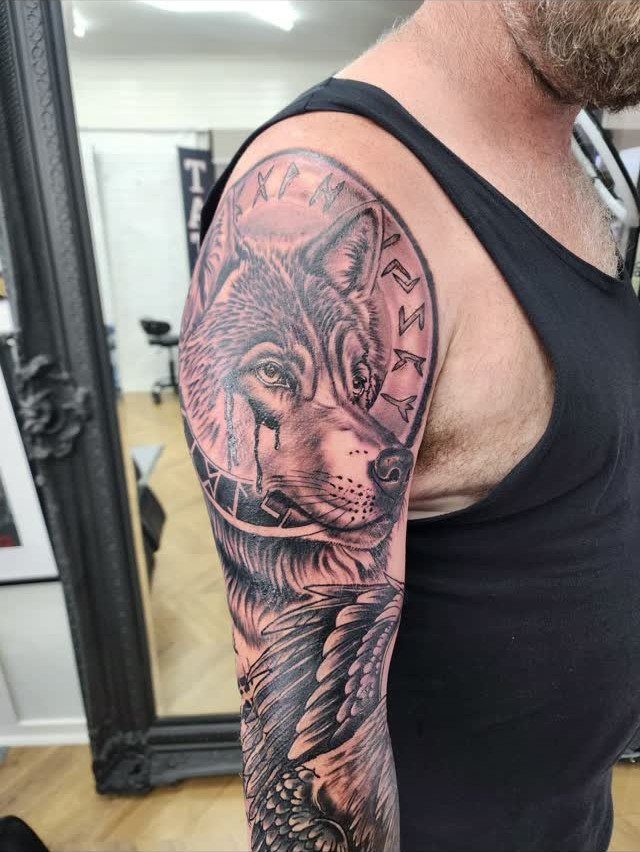 Nordic Runes Tattoo: 10 Design Ideas Worth Considering 8 A detailed black and grey wolf tattoo with Nordic runes on a man's arm.