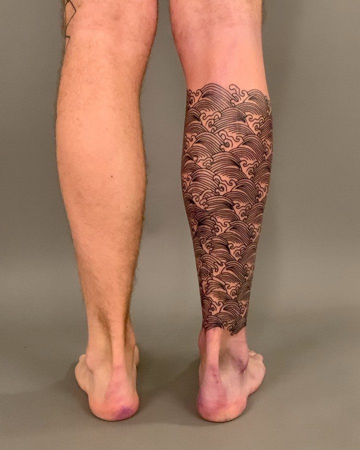 A person's lower legs featuring a large black ink tattoo of repetitive ocean wave patterns on the right calf.