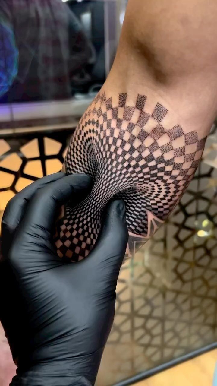 A black ink optical illusion tattoo on a forearm creating a distorted checkerboard effect.