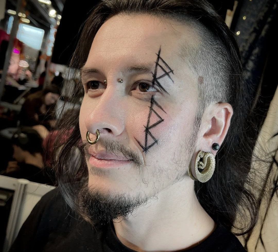Rune Tattoo Ideas: 18 Symbols and Placements Worth Studying 12 A man with a black geometric rune tattoo on his cheek and facial piercings.