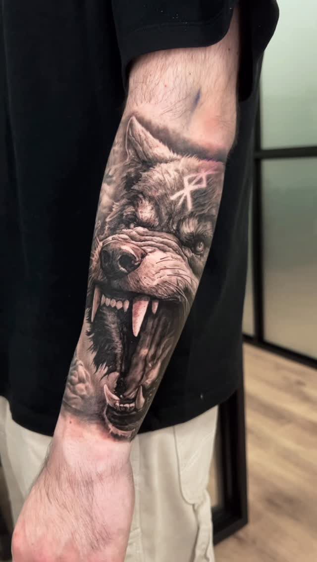 Rune Tattoo Ideas: 18 Symbols and Placements Worth Studying 8 A detailed black and grey wolf head tattoo with a rune symbol on a person's forearm.
