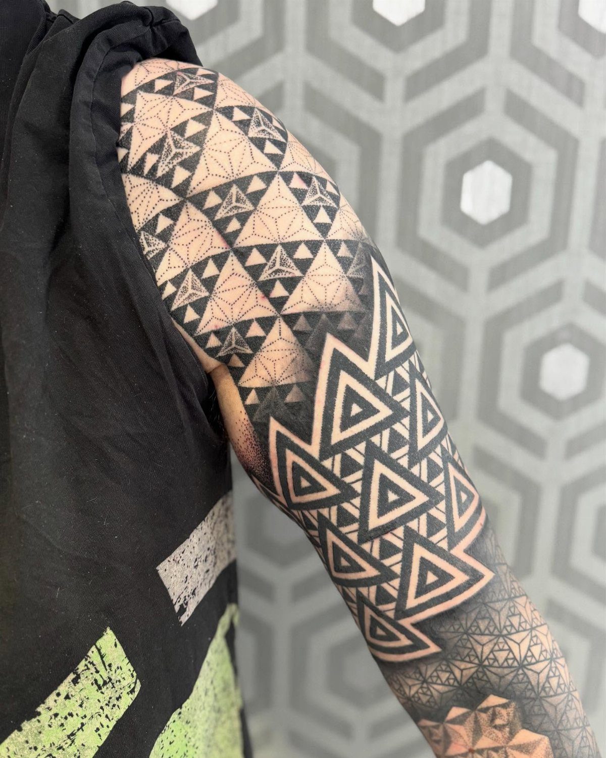 A person's arm covered in a large, intricate black ink geometric tattoo featuring many small triangles.
